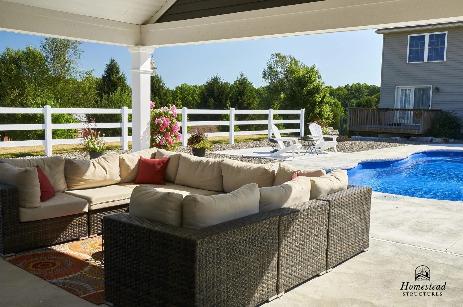 Covered outdoor patio with beige sectional sofa with red pillows, next to a swimming pool with a concrete deck, surrounded by a white wooden fence, with green trees and a house in the background.