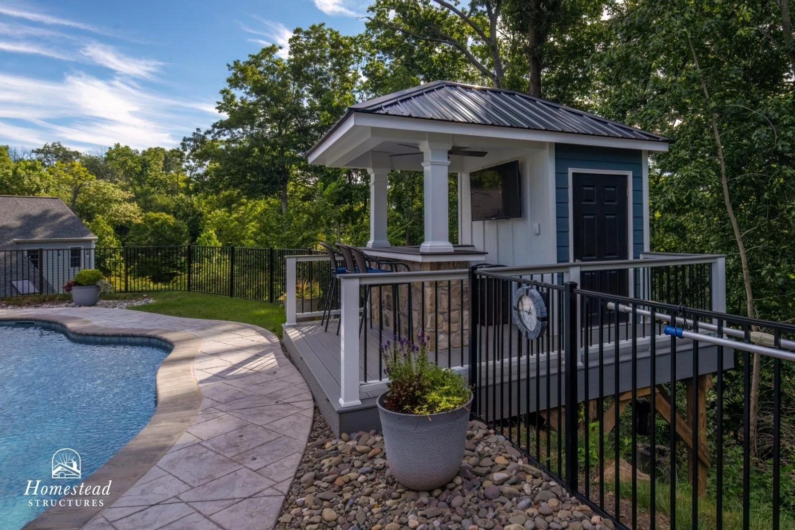 A backyard pool area featuring a pool with a curved edge, a small elevated pool house with a TV, black door, and a roof supported by columns, surrounded by trees and a fence.