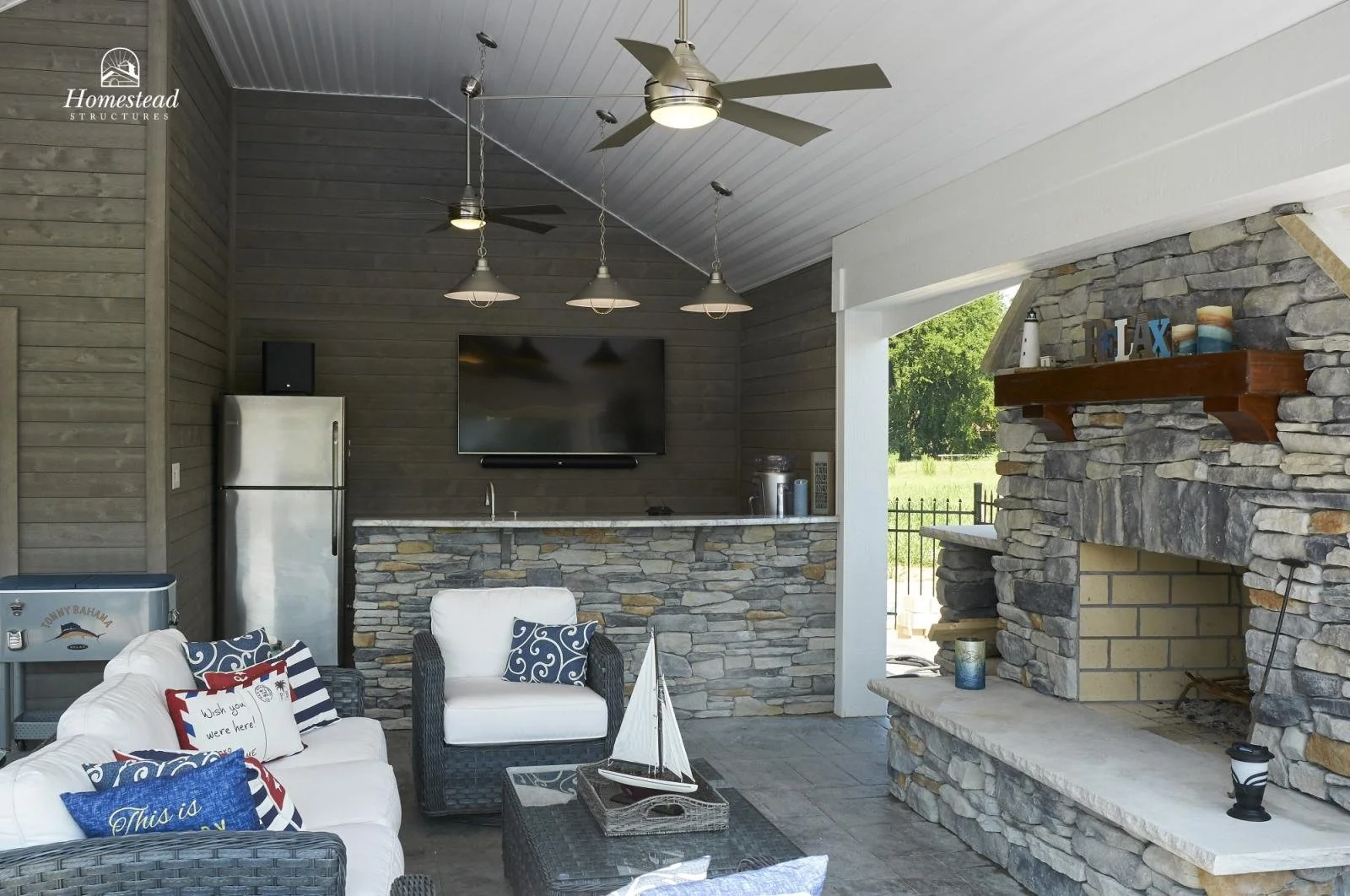 Outdoor living space with stone fireplace, ceiling fan, flat-screen TV, and furniture including a sofa and chairs with nautical-themed pillows.