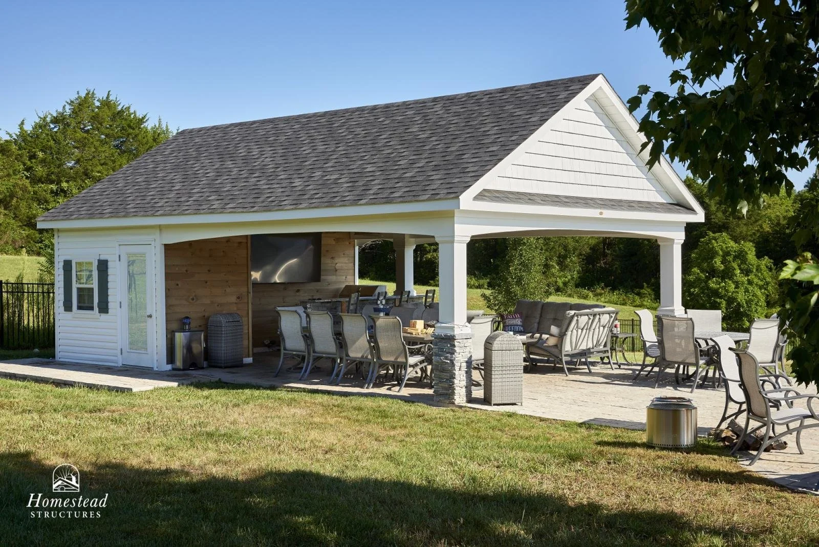 An outdoor pavilion with white siding, gray shingle roof, and stone accents, furnished with patio tables, chairs, and a sofa, set in a grassy backyard with trees.