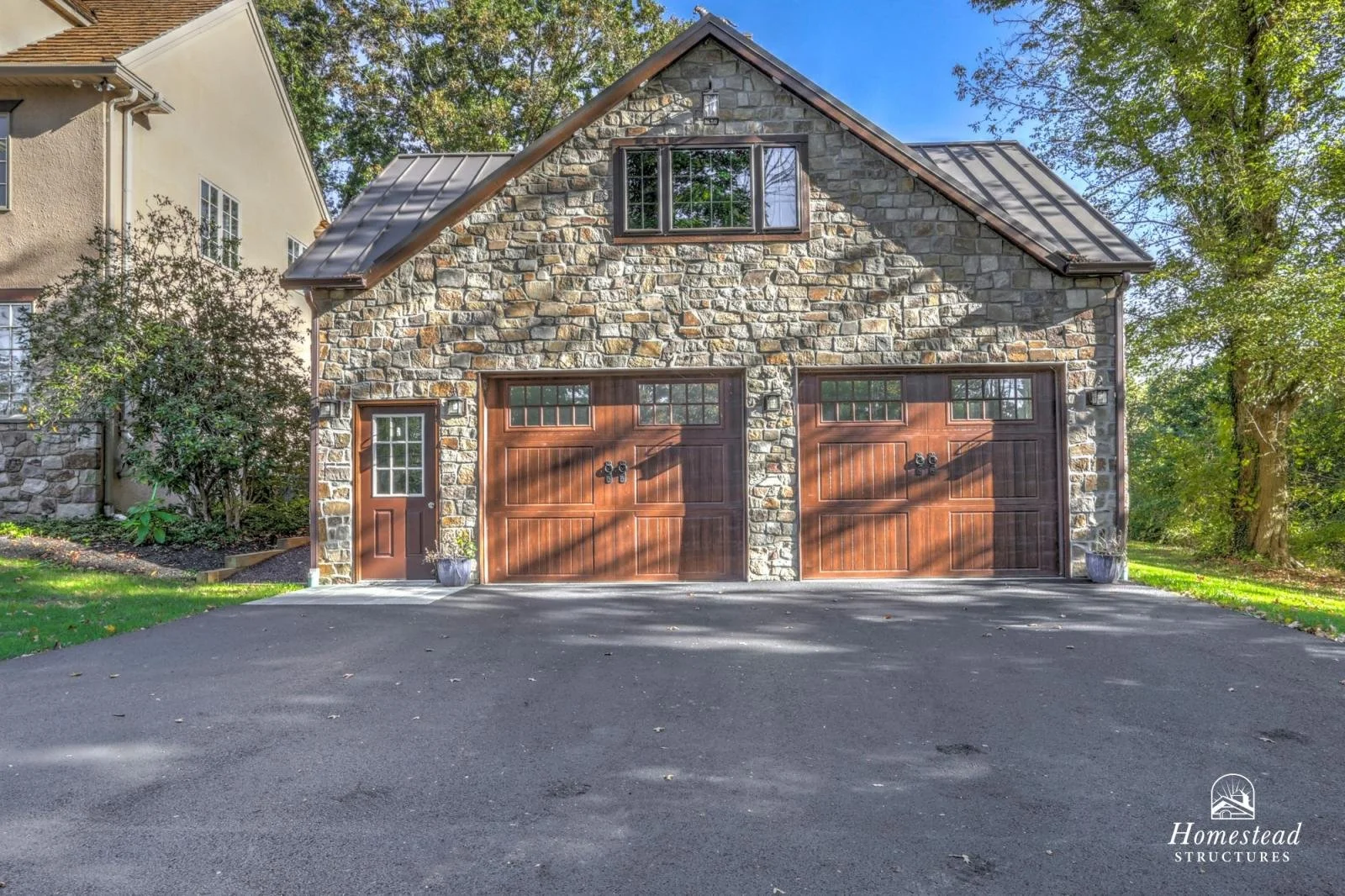 Front view of a house with a stone facade, two wooden garage doors, a side door, and a pitched roof, surrounded by trees and a paved driveway.