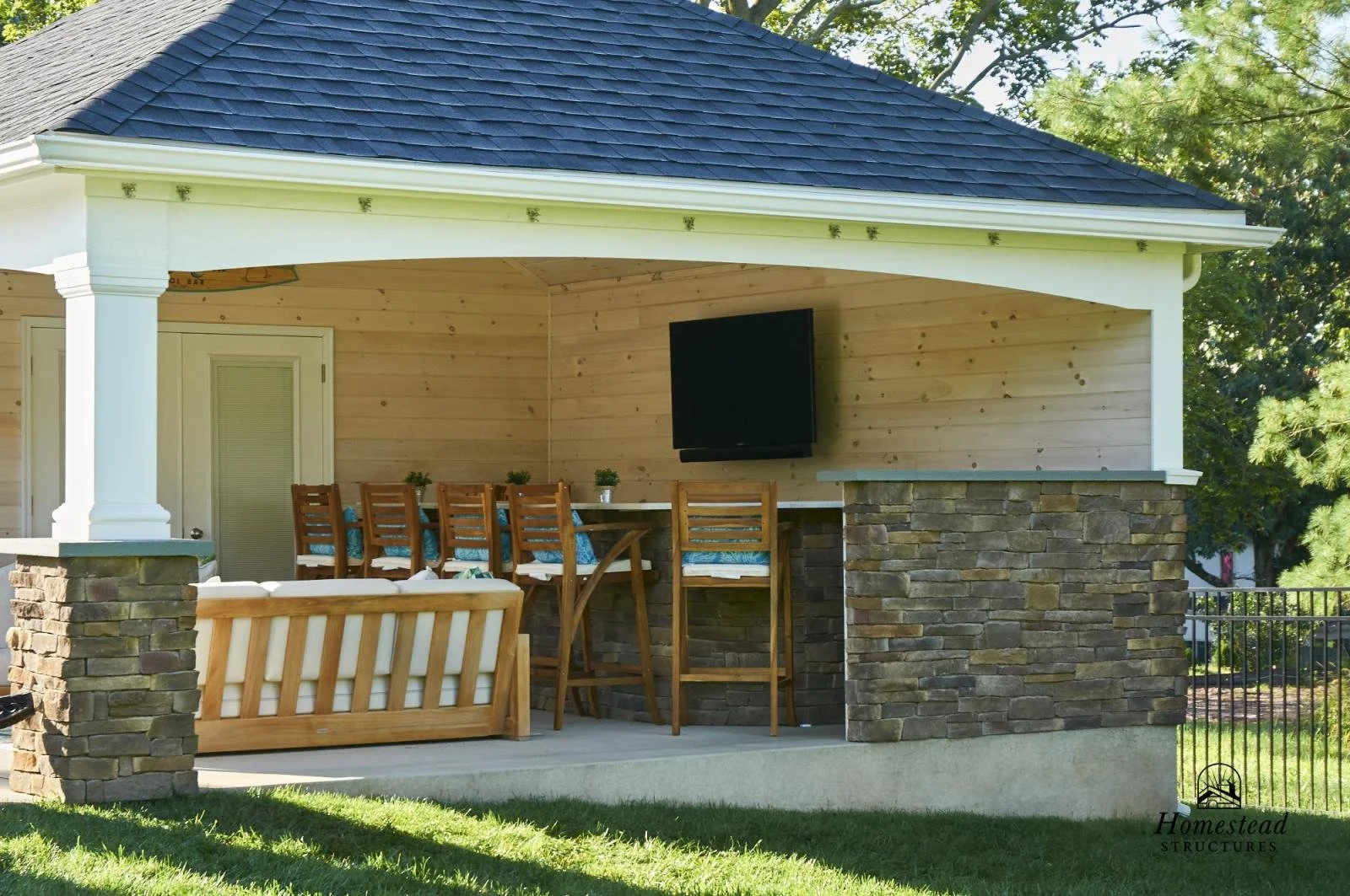 Outdoor covered porch with wooden and stone accents, featuring a mounted TV, a long wooden table with chairs, and a cushioned wooden bench, surrounded by a grassy lawn and trees.