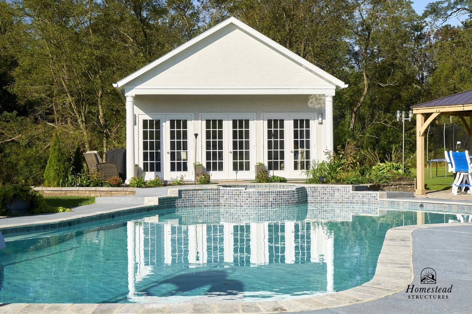 A backyard with a swimming pool in front of a white pool house with glass doors and columns. There is outdoor furniture and a grassy area with trees in the background.