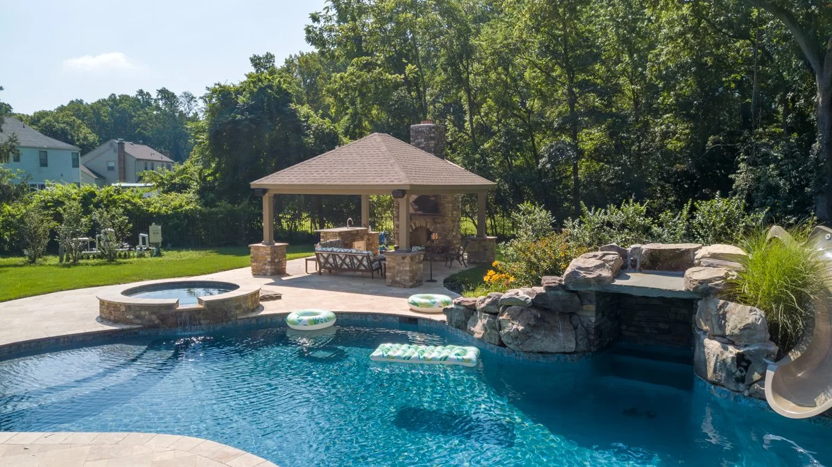 Backyard with a swimming pool, hot tub, rock waterfall, gazebo with seating, and surrounding trees and houses.
