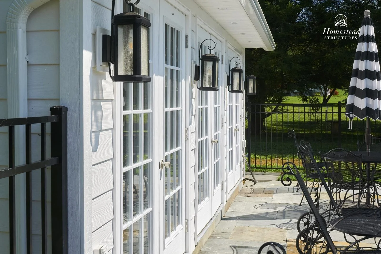 Exterior view of a house with white French doors and multiple black outdoor wall lanterns. A patio with wrought iron furniture, a striped patio umbrella, and a fenced yard with green grass and trees in the background.