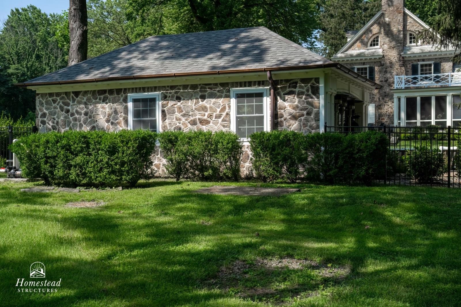 A stone cottage with a gray shingled roof, two white-framed windows, and a front lawn with trimmed bushes and green grass.