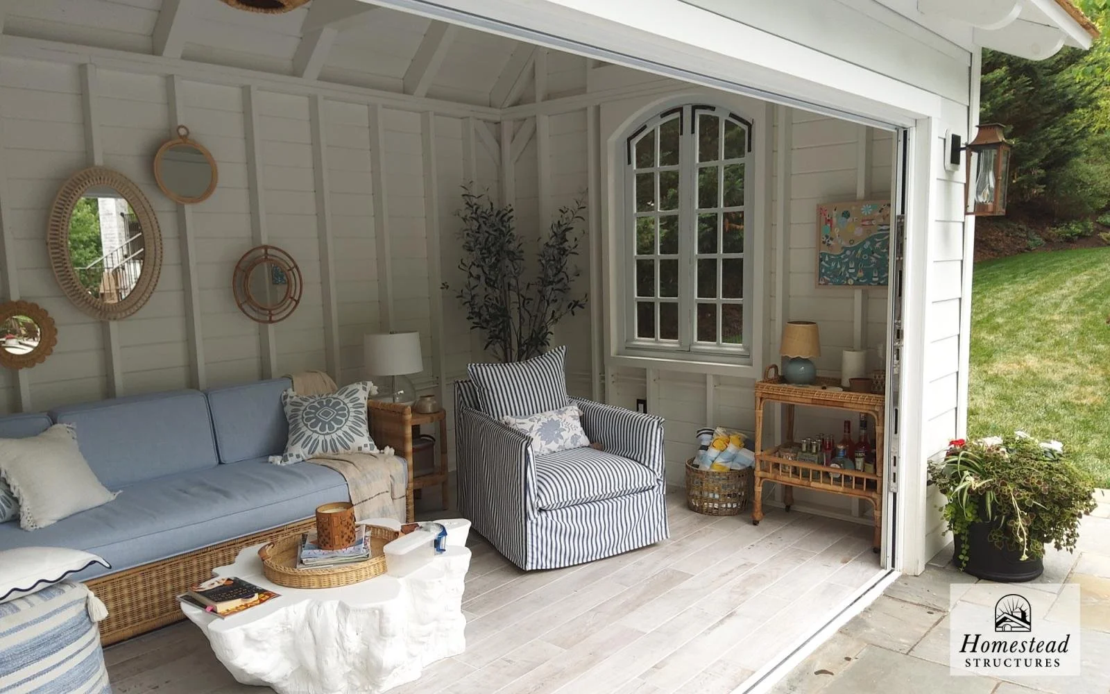 Cozy sunroom with white paneled walls, a blue sofa, a striped armchair, and various decorative mirrors on the wall, with outdoor scenery visible through a window and open door.