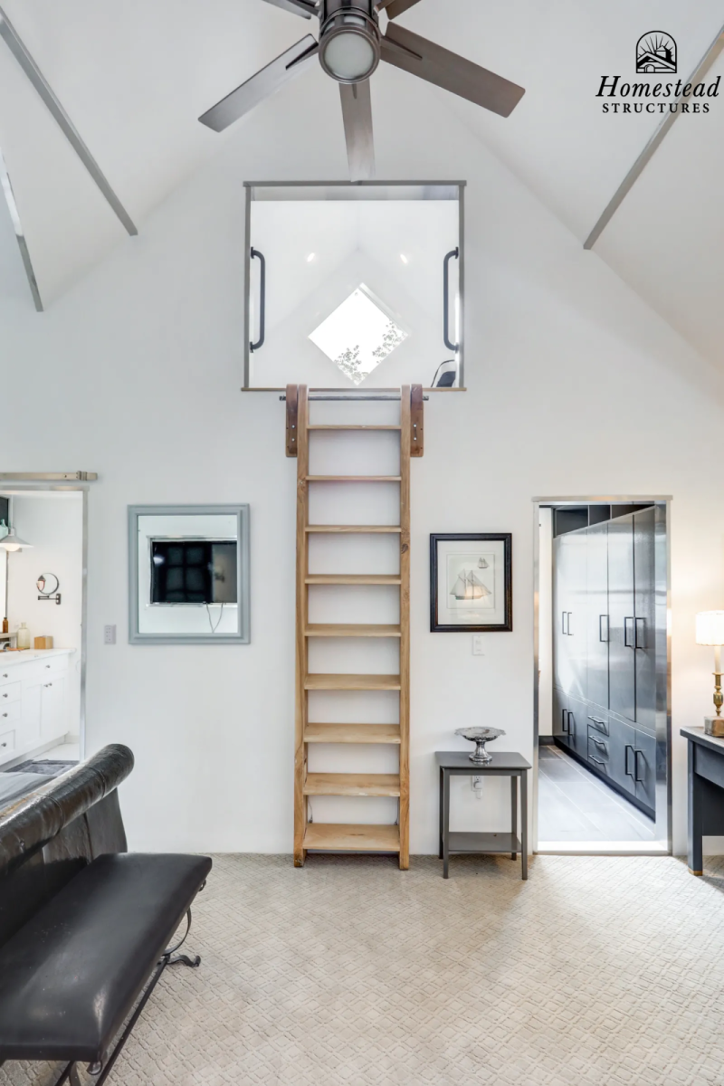Living room with ladder leading up to a square opening in the ceiling with a window. Ceiling fan overhead, wall-mounted TV, framed artwork, small side table, and black storage cabinet.