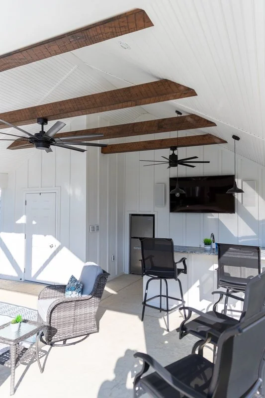 Outdoor patio with wicker chair, glass-top table, and black chairs near a white wall with a mounted flat-screen TV, ceiling fans, and wooden beams on the ceiling.