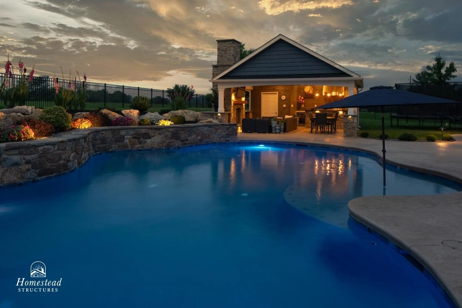 A backyard with a swimming pool at dusk, a stone wall with colorful flowers, a covered outdoor living area with string lights, a large patio umbrella, and a grassy yard with a fence in the background.