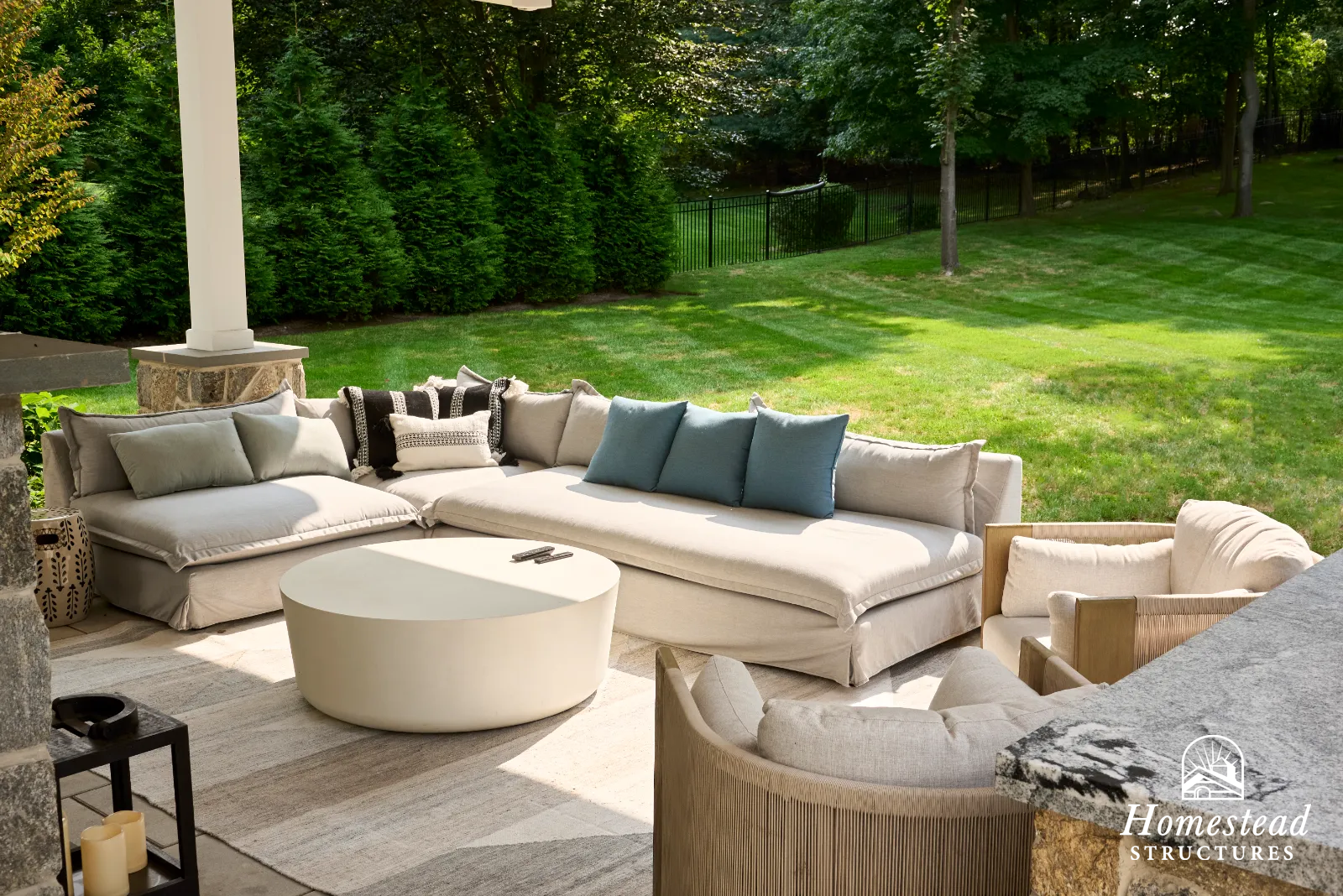 Outdoor patio with beige sectional sofa, blue and patterned cushions, white round coffee table, armchair, and view of green lawn and trees in the background.
