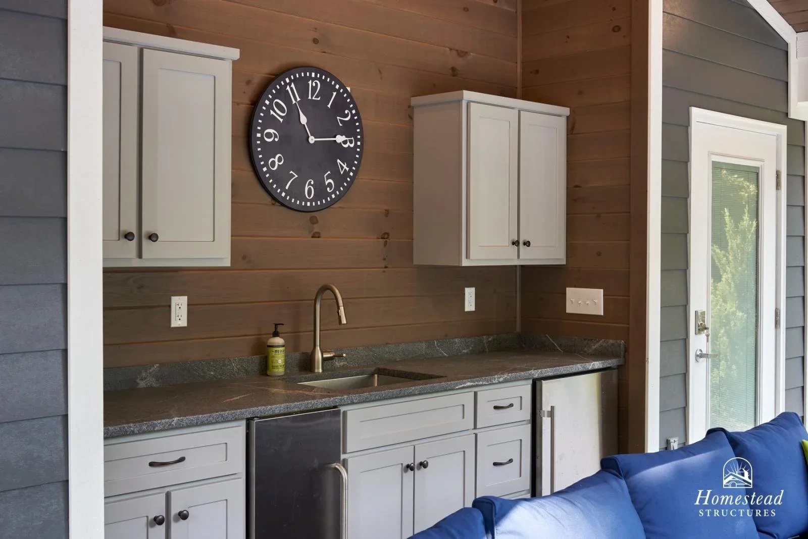 Interior of a small kitchen or kitchenette with a wooden wall, white cabinets, a black wall clock showing 11:53, a sink with a gold faucet, a soap dispenser, and a granite countertop. There is an outdoor door with a glass window on the right side.