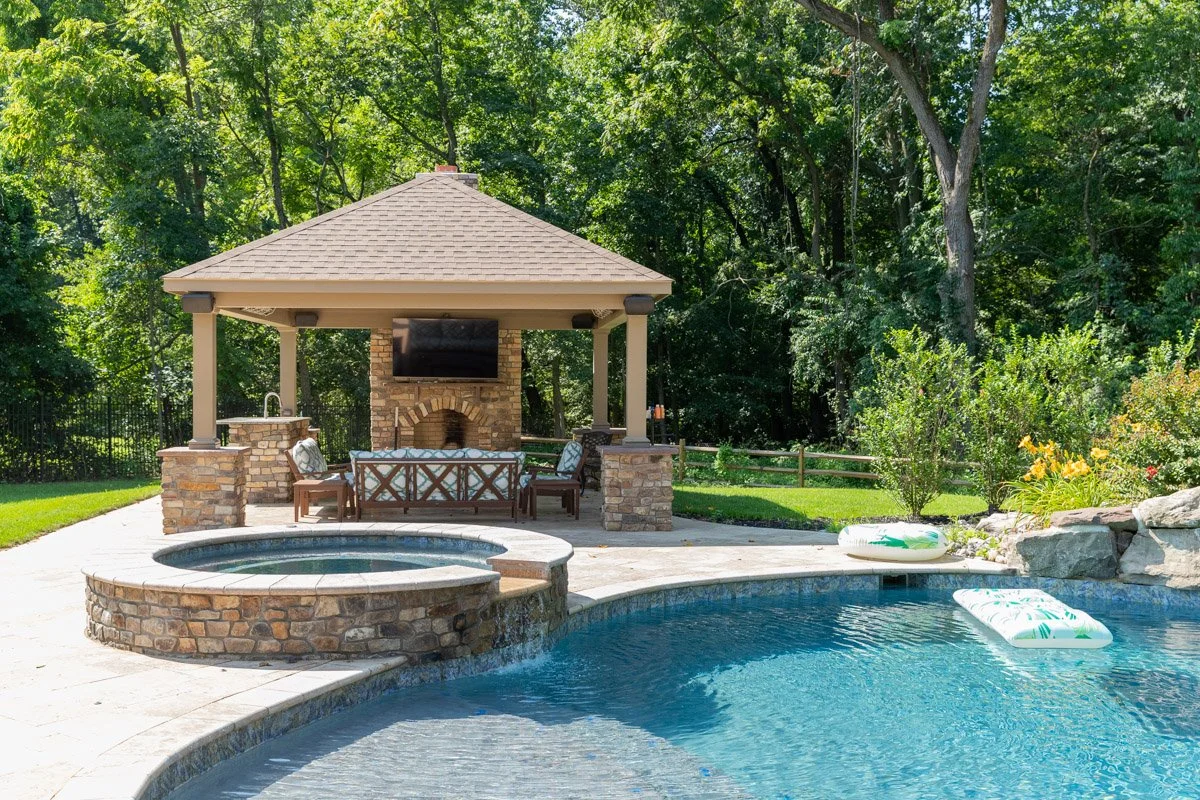 Backyard swimming pool with a hot tub in a landscaped yard. There is a pavilion with a television, seating, and a fireplace in the background, surrounded by trees and greenery.