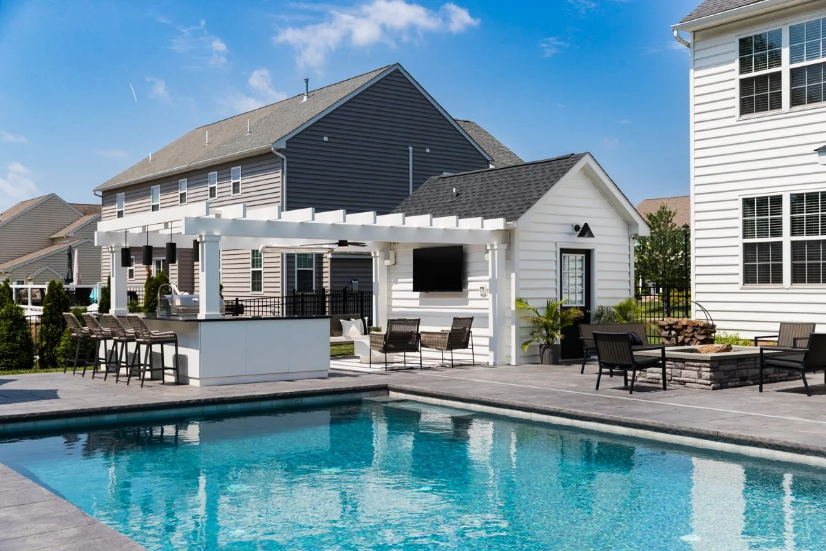 Backyard with swimming pool, outdoor seating, a white pool house with a mounted TV, and a barbecue area with a counter and bar stools, surrounded by suburban houses.