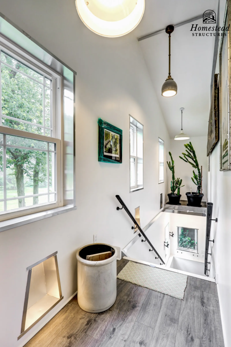 Bright hallway with large windows, potted cacti, artwork on the walls, a ceiling light, and a staircase with a black handrail.