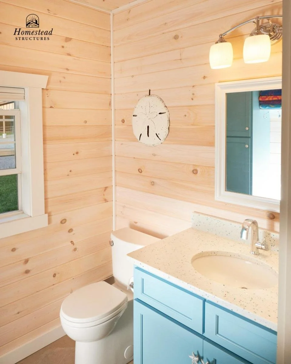 Small bathroom with light-colored wooden walls, a white toilet, a blue vanity with a speckled countertop, a mirror, and a window revealing a grassy outdoor area.