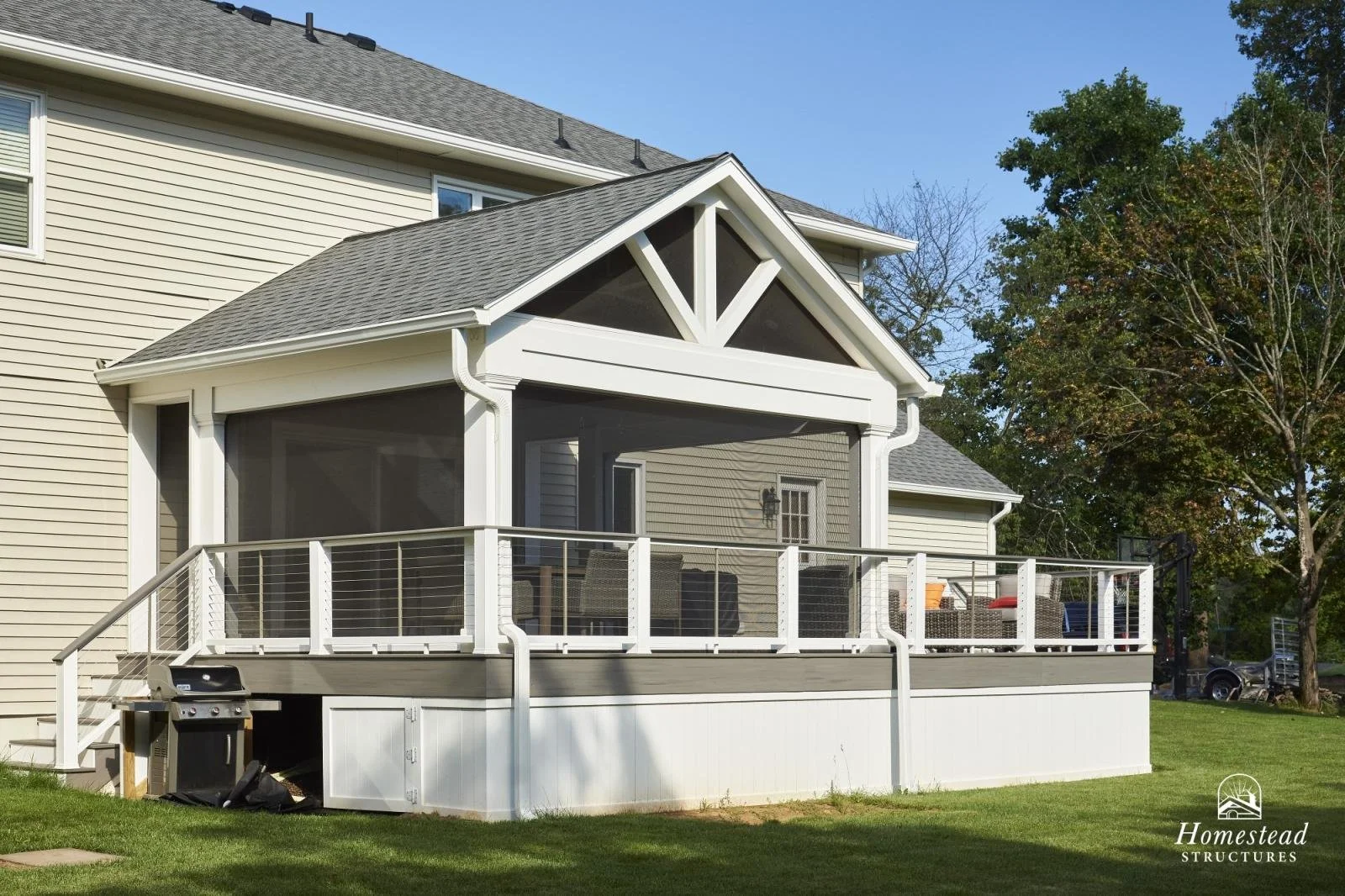 A house with a white covered porch, screened-in sides, and outdoor furniture including chairs and a small table, surrounded by green yard and trees.