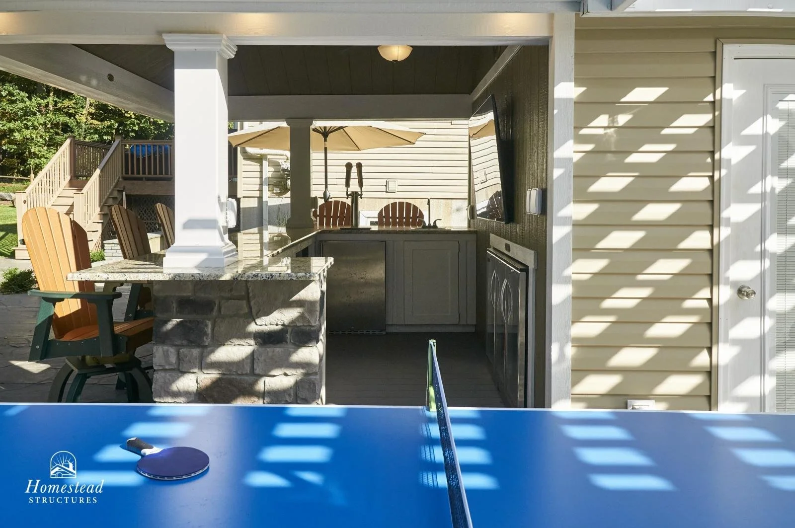 View of an outdoor covered patio area with a ping pong table in the foreground, a bar area with a stone counter, a refrigerator, a sink, and chairs under a large umbrella in the background, with a staircase and greenery to the left.