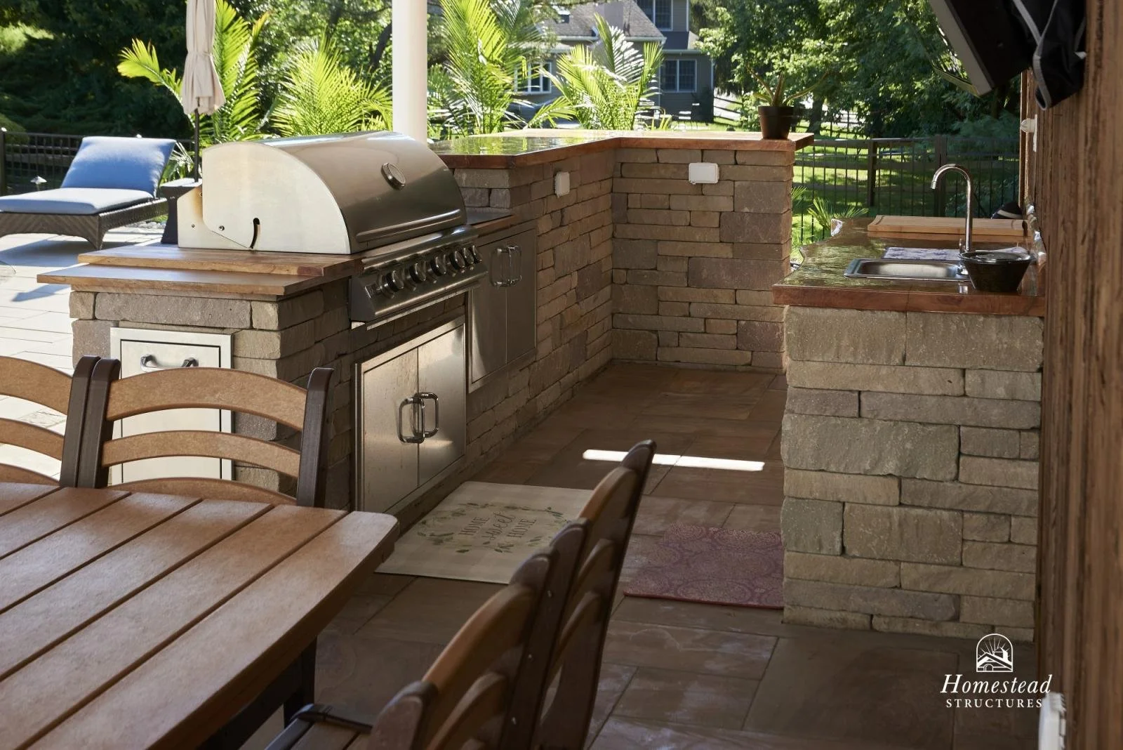 Outdoor kitchen with brick and stone counter, stainless steel grill, sink, and outdoor seating area with wooden table and chairs, surrounded by greenery.