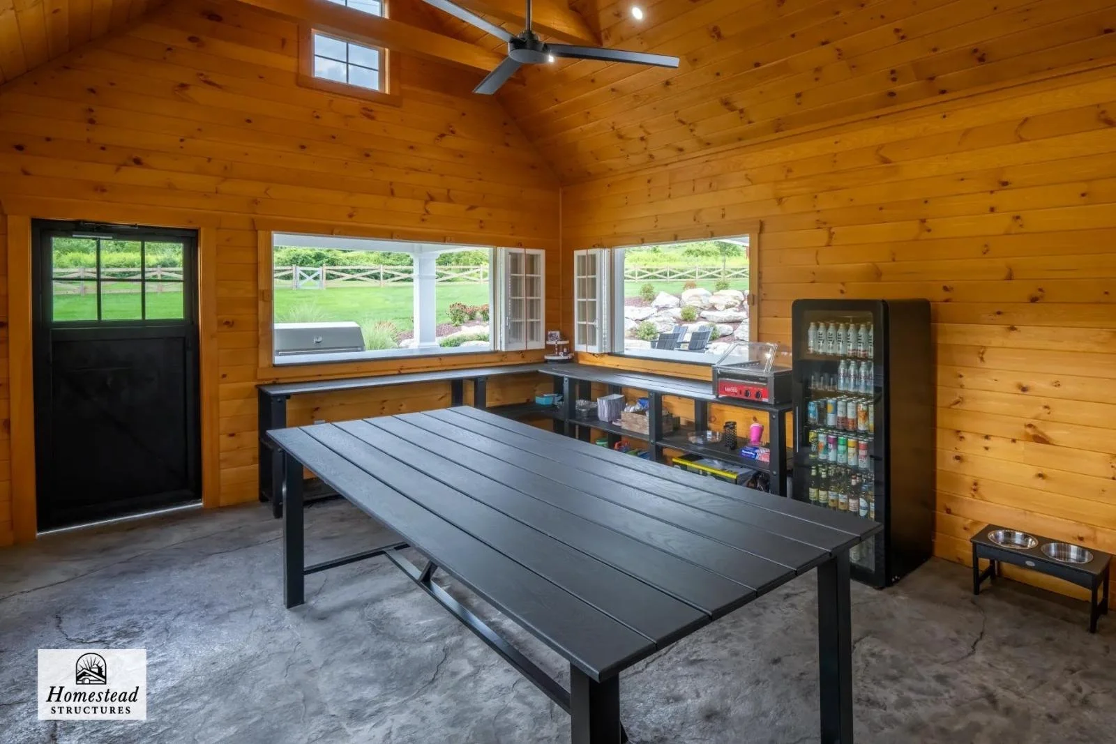 Interior of a wooden outdoor pavilion with large windows, a black table, a black beverage refrigerator, and a pet feeding station, overlooking a landscaped yard.