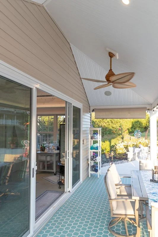 Covered outdoor patio area with sliding glass doors leading inside. There is a ceiling fan, a white shelving unit with towels, a table with chairs, and a view of a backyard with trees and garden decorations.