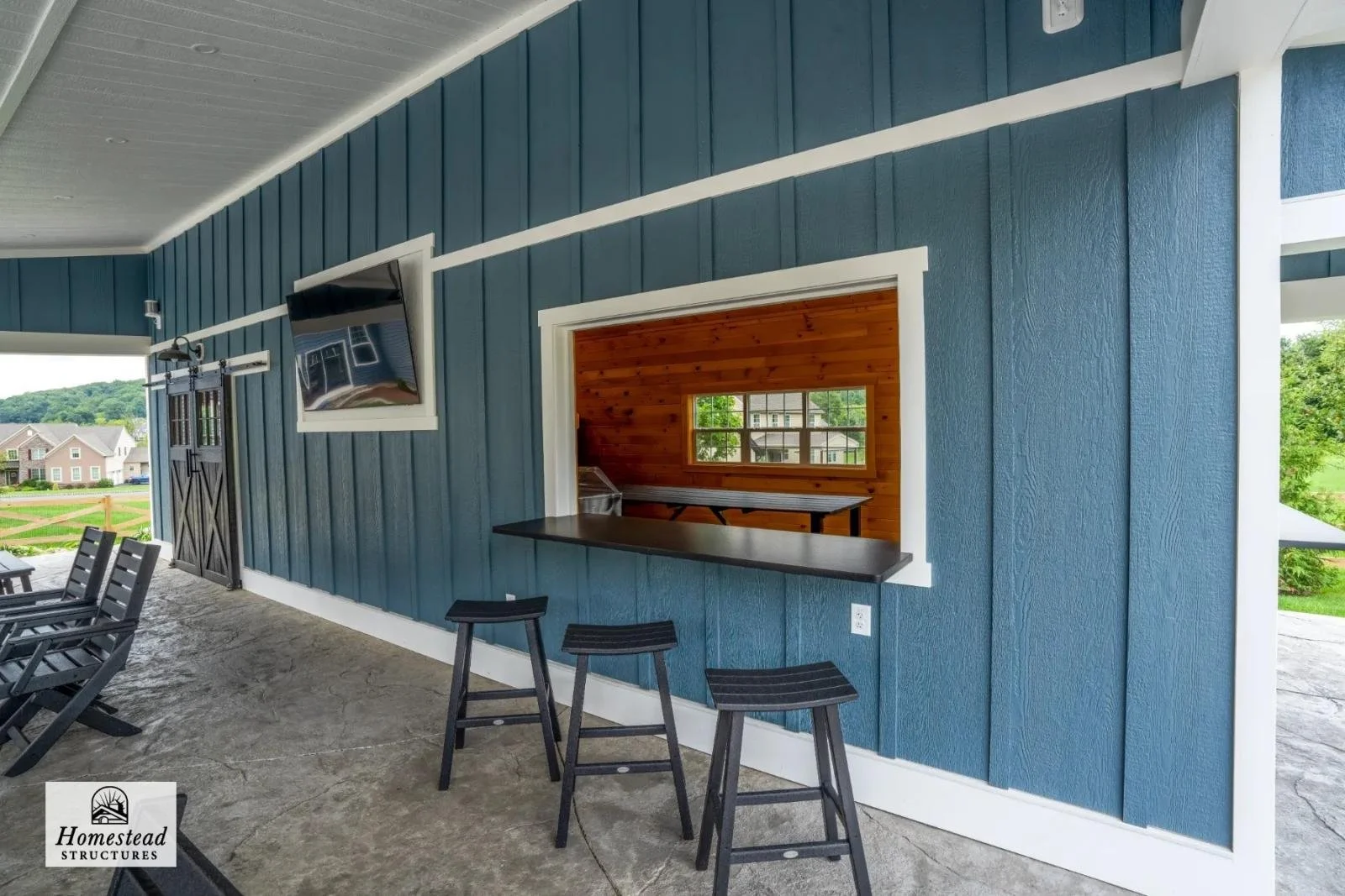 Outdoor covered patio area with a blue wooden wall, mounted flat-screen TV, and a serving window with a black countertop, three black barstools, and a view of neighboring houses and greenery.