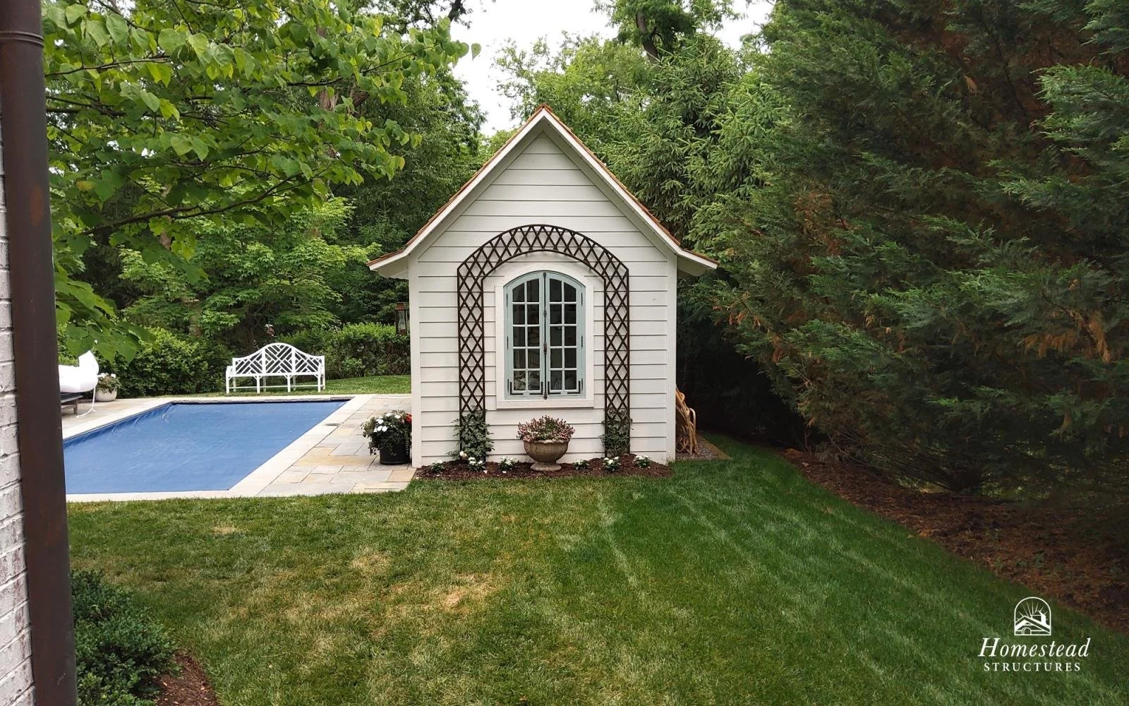 A small white garden shed with window and decorative brown trellis in a backyard with a swimming pool, green lawn, trees, and bushes.