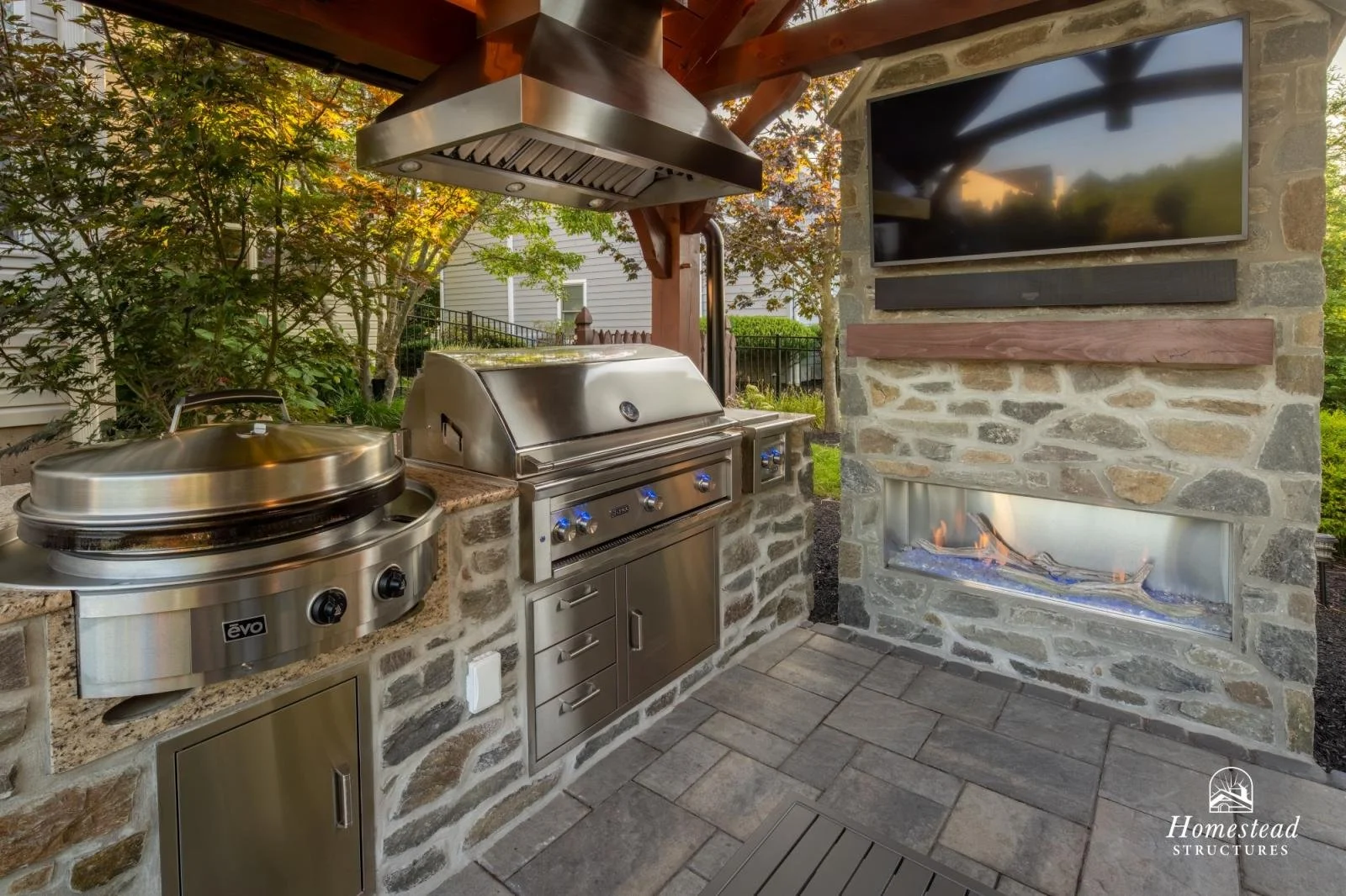 Outdoor kitchen with stainless steel grill, a warm fire in a built-in stone fireplace, a large TV mounted on a stone wall, and surrounding greenery.