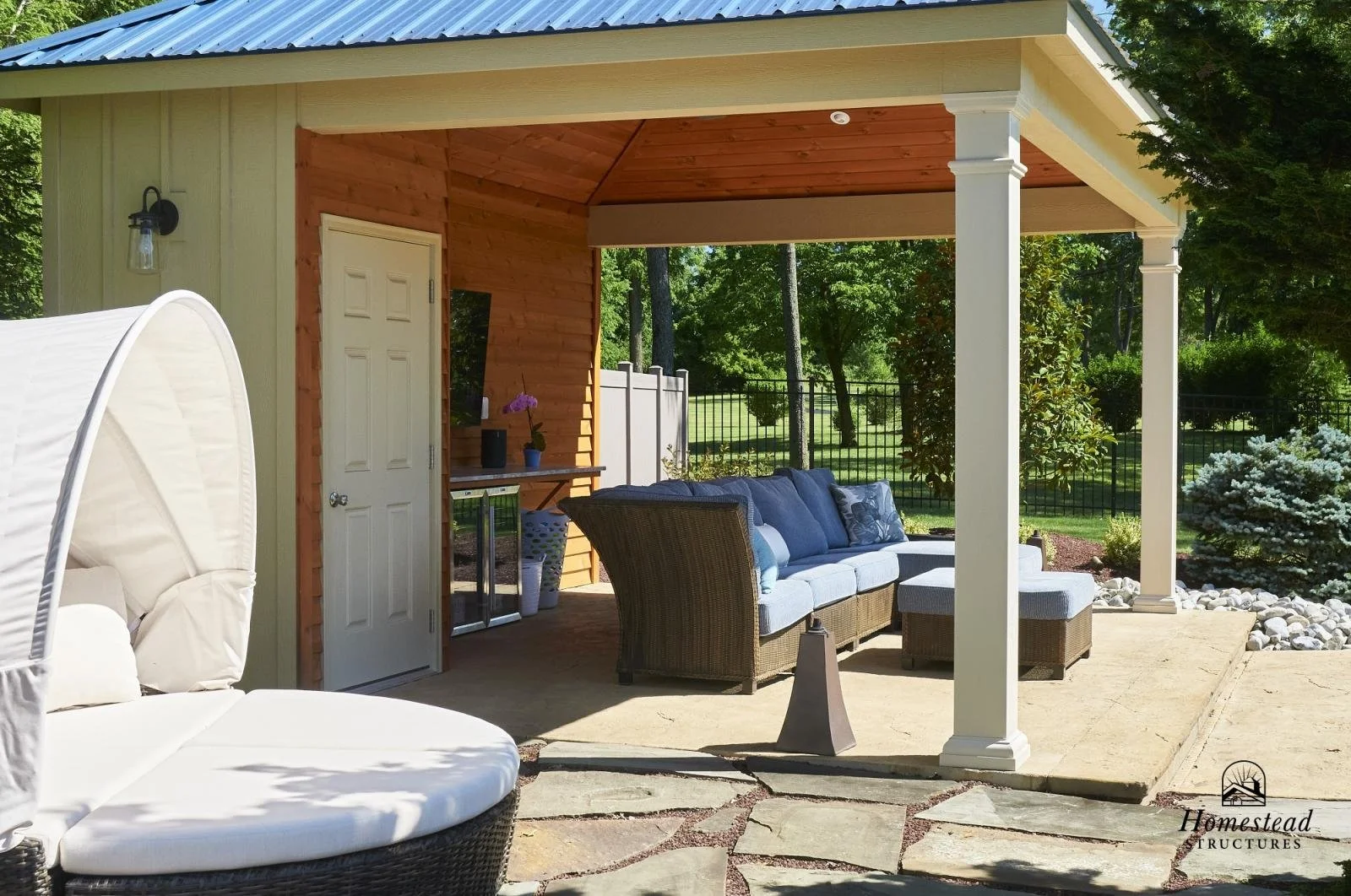 Covered outdoor patio with wicker sofa, chairs, and a potted plant, surrounded by trees and a black fence.