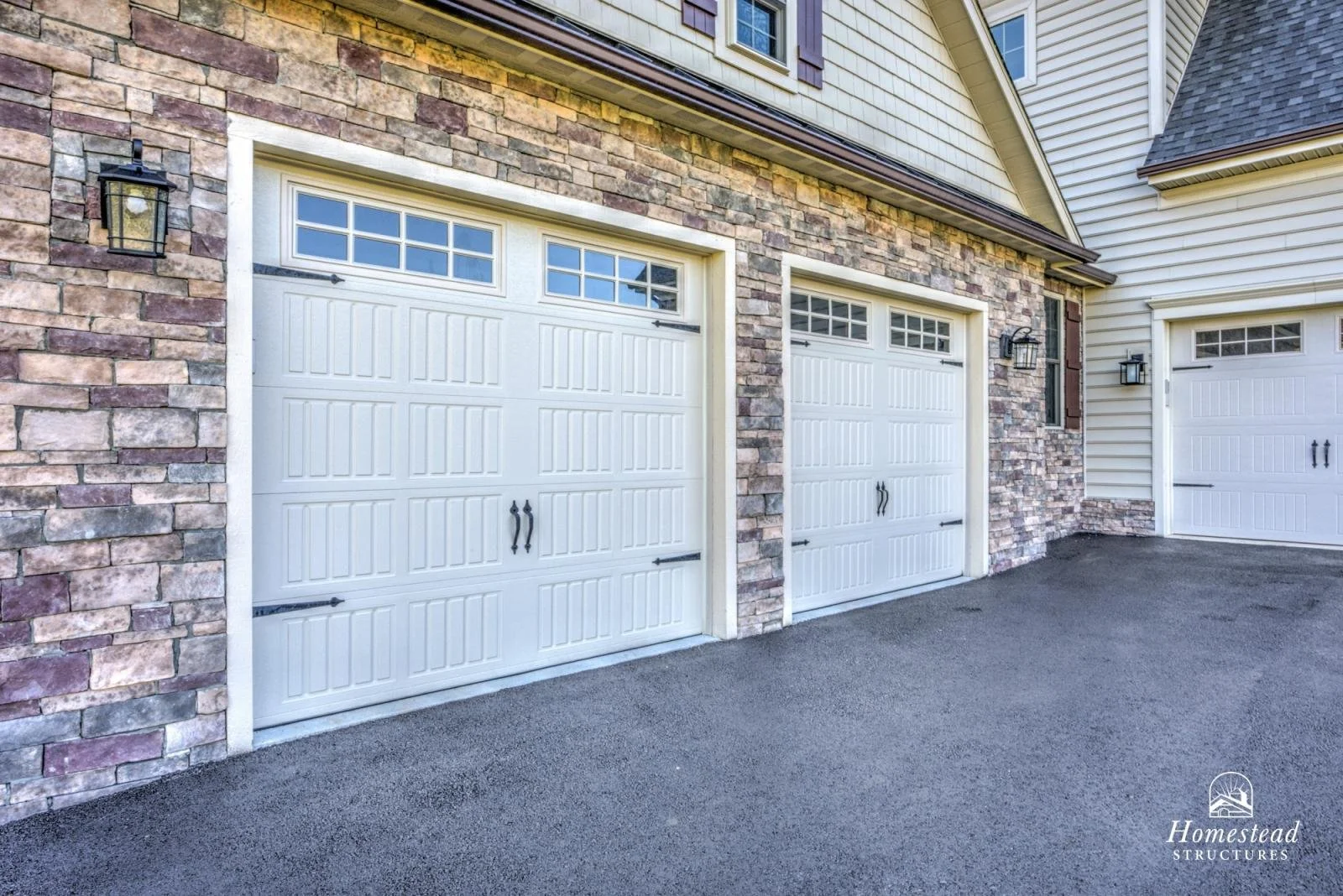 Three white garage doors with windows at the top, attached to a building with stone and siding exterior.
Black exterior lights are positioned on the wall near each garage door.
The ground in front is paved with asphalt.