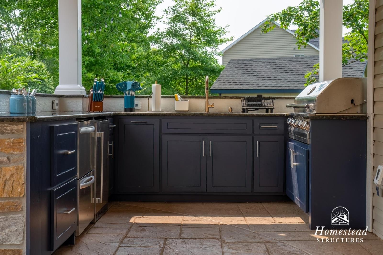 Outdoor kitchen area with dark cabinets, a countertop, a grill, and various kitchen utensils, with a view of trees and neighboring houses.