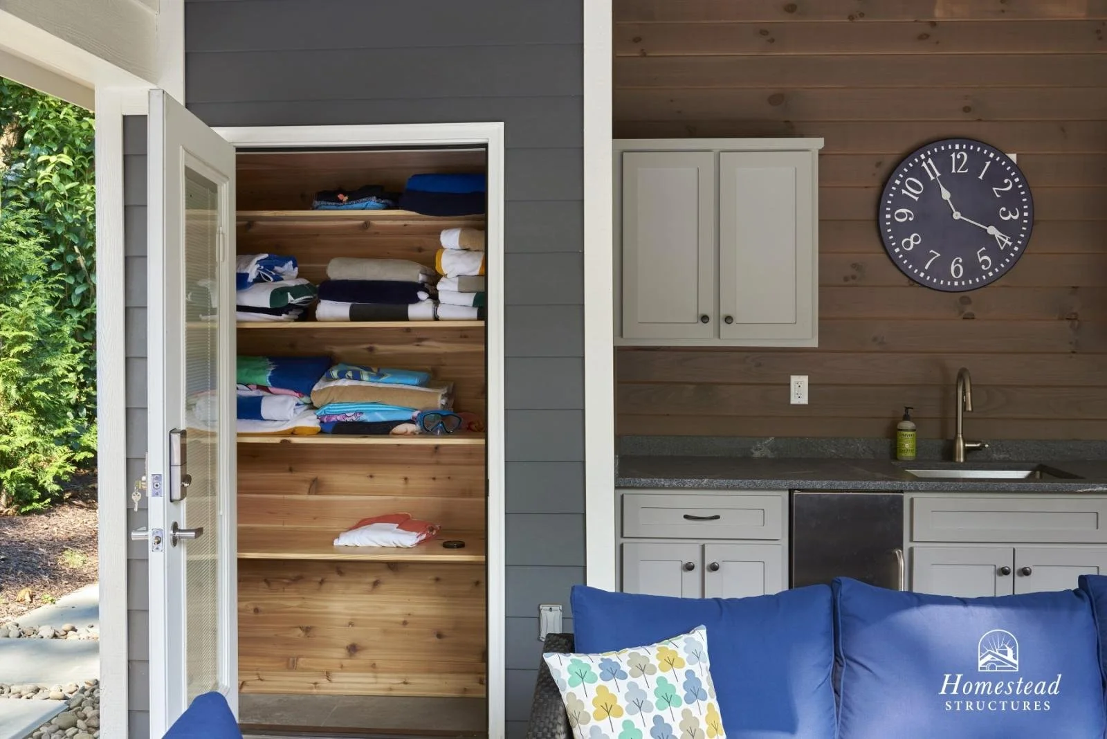 Indoor view of a small kitchen area with a wall clock, white cabinets, a sink with a bottle of soap, and a navy blue sofa with colorful pillows. An open door reveals shelves of folded clothing and towels.