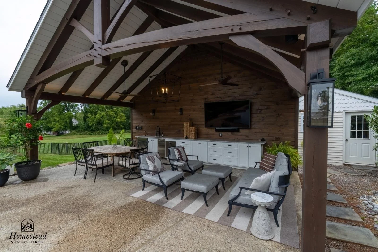 Outdoor covered patio with seating area, including a sofa, armchairs, and a rug. There is a built-in white cabinet, a wall-mounted TV, and ceiling fans. The patio has a wooden roof structure with exposed beams.