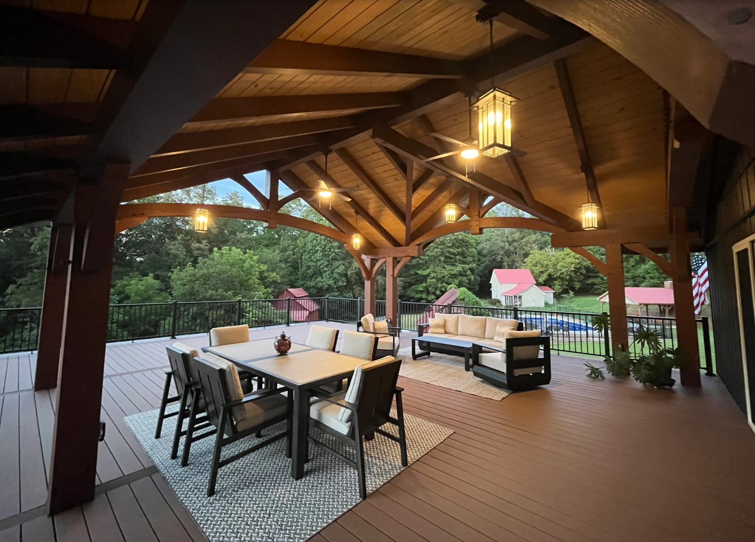 Covered outdoor patio with wooden ceiling, hanging lights, outdoor furniture including a dining table with chairs, a corner sofa, and potted plants, overlooking a green landscape with trees, sheds, and American flag on the right side.