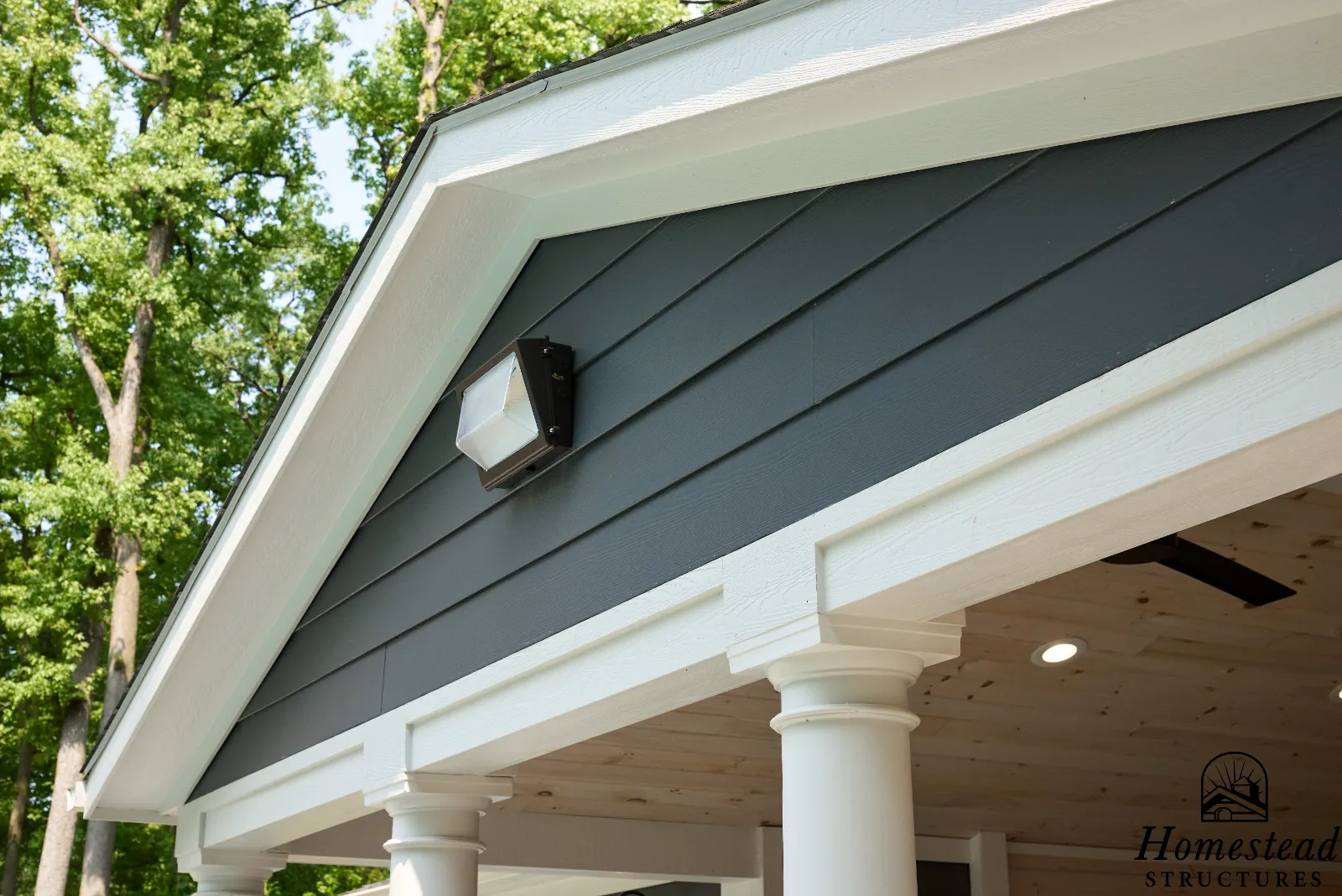 Close-up of the front porch of a house with black siding, white trim, columns, outdoor lighting, and ceiling lights, surrounded by green trees.