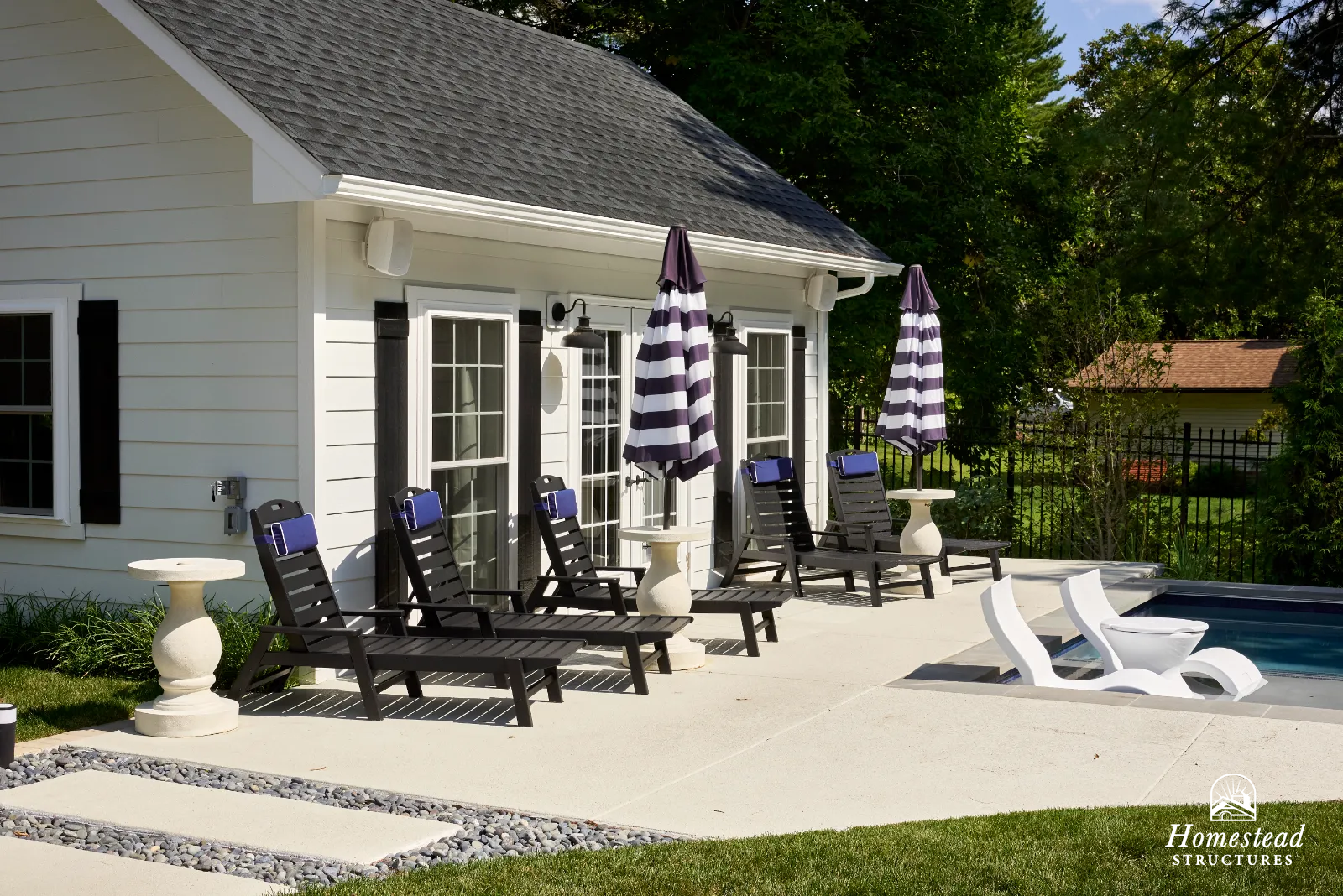Backyard patio area with four black lounge chairs with purple accents, two black and white striped umbrellas, a small side table, and a modern white lounge chair near a swimming pool, with a white house and green trees in the background.