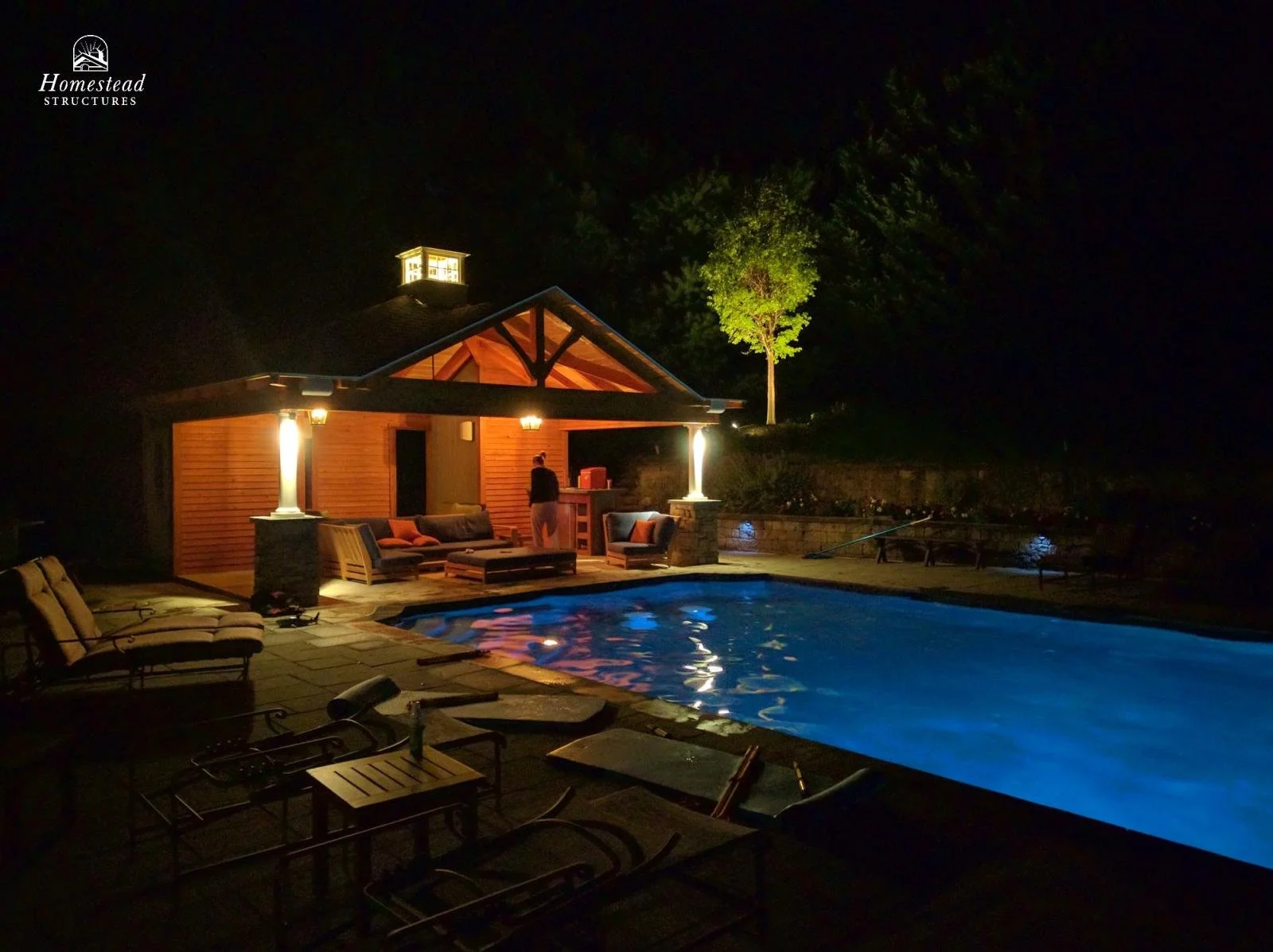 Night view of a backyard with a lit swimming pool, outdoor seating area, a small building, and a lit tree, with a person standing near the building.