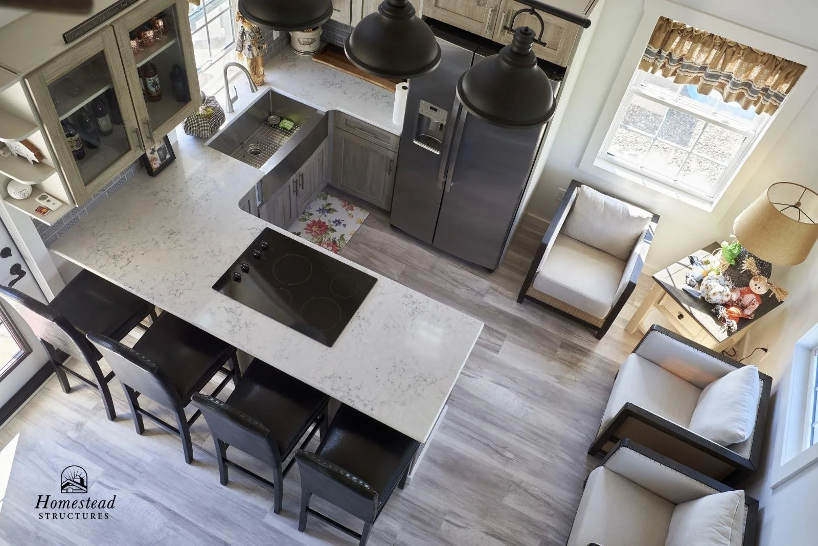 A cozy kitchen and sitting area viewed from above, featuring a white countertop with black chairs, a stainless steel refrigerator, a sink, a stove, a window with plaid curtains, an armchair, a small side table with a lamp, and a white armchair, decor