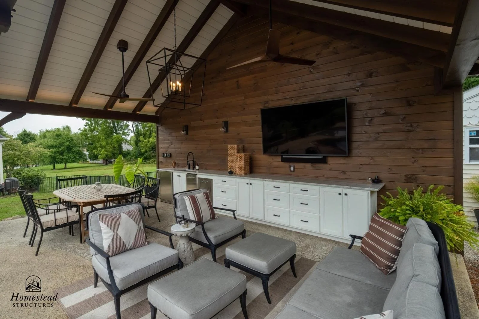 Covered outdoor patio with a white kitchen counter, chairs, and a large flat-screen TV mounted on a wooden wall, overlooking a green yard.