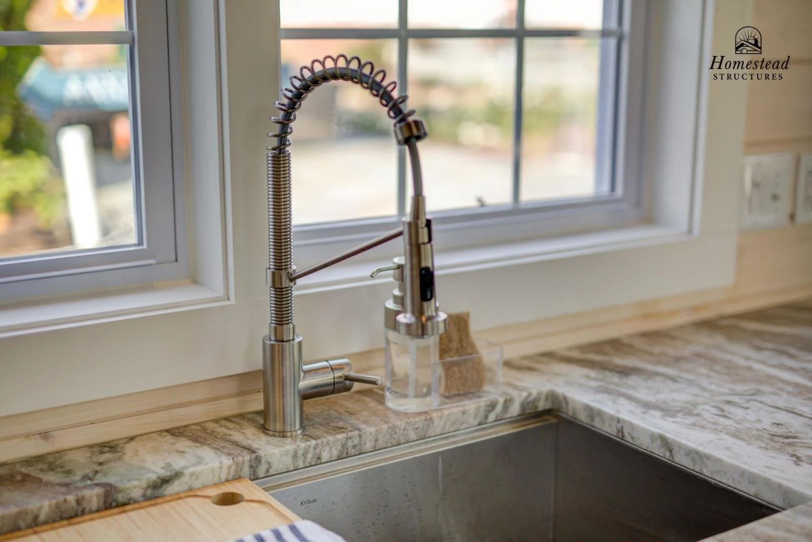 Kitchen sink with stainless steel faucet and spring coil neck, situated under a window with a view of a porch or yard area. The countertop is made of granite or marble, and a sponge is placed near the sink.