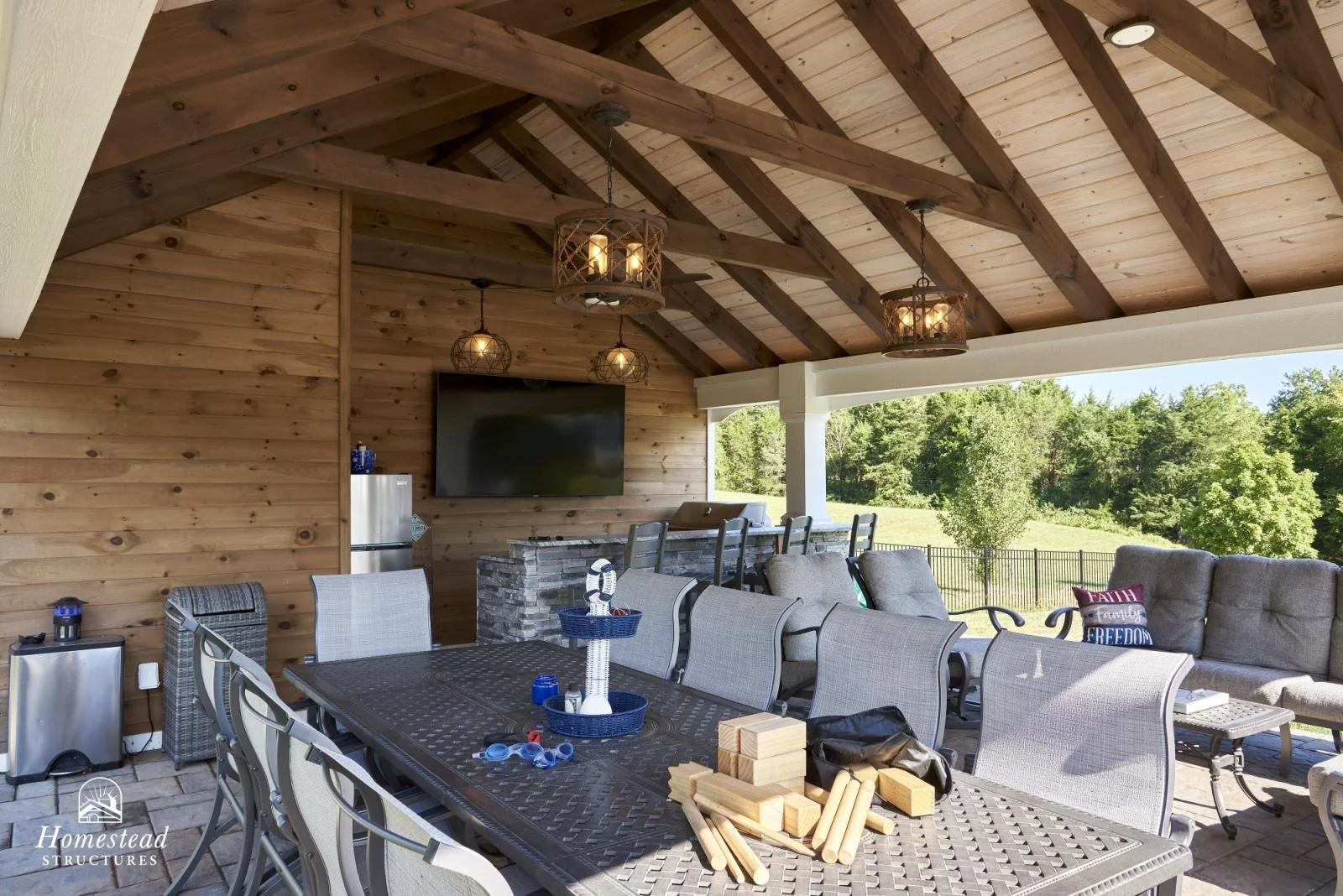 Covered outdoor patio with wooden ceiling and walls, a large dining table with chairs, a lounging area with cushioned chairs, a TV on the wall, and outdoor scenery with trees and grass.