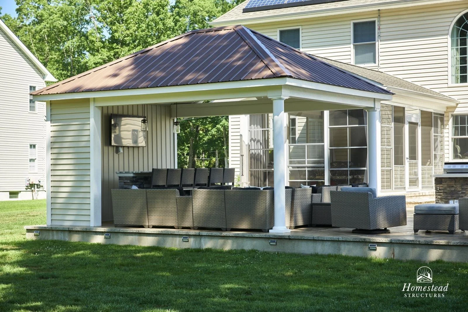 A backyard patio with outdoor seating under a gazebo, with a television mounted on one side and a grill in the corner. The area is surrounded by green grass and neighboring houses.