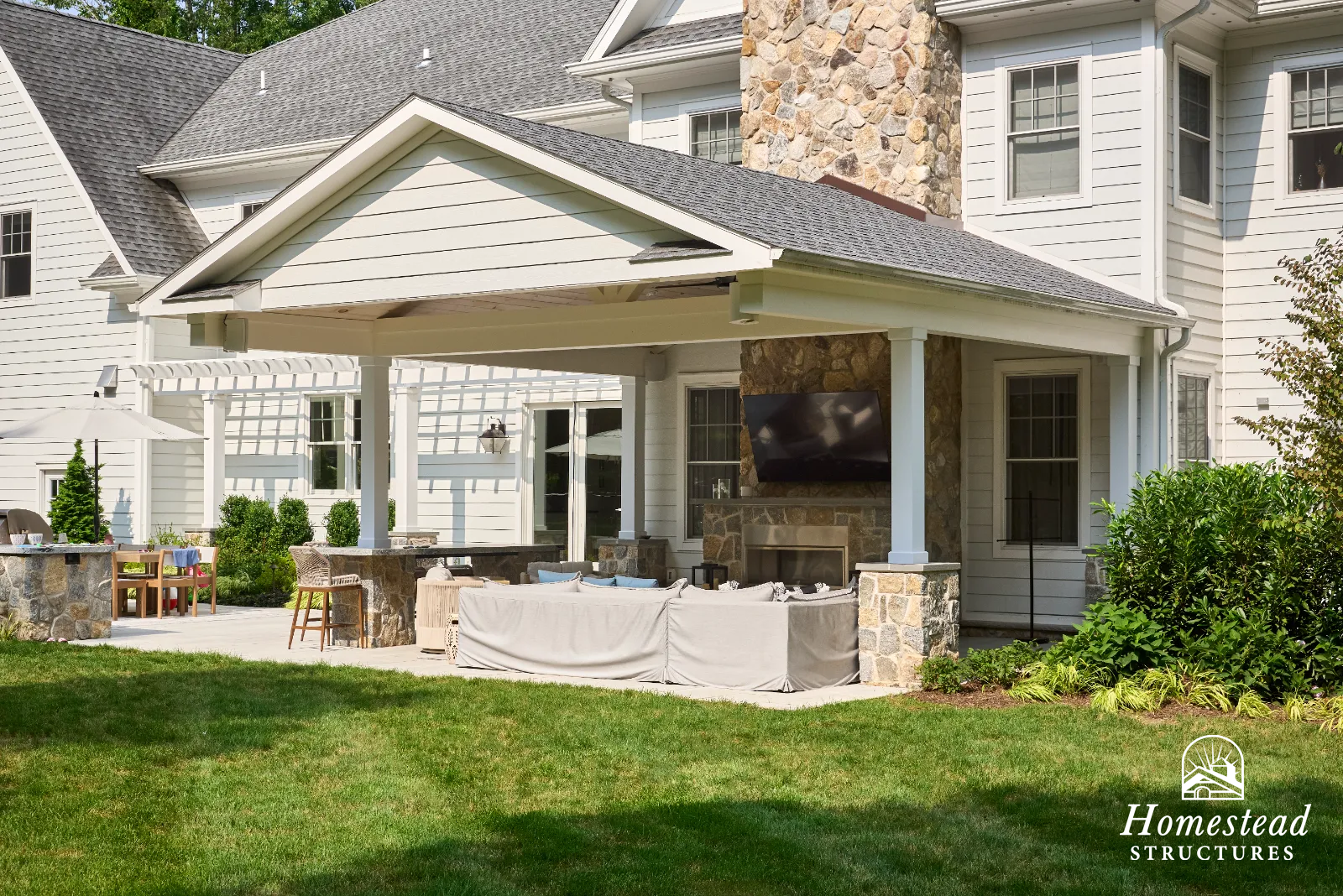 A backyard patio area with outdoor furniture, a stone fireplace, and a mounted television under a white pergola attached to a large house with white siding and multiple windows.