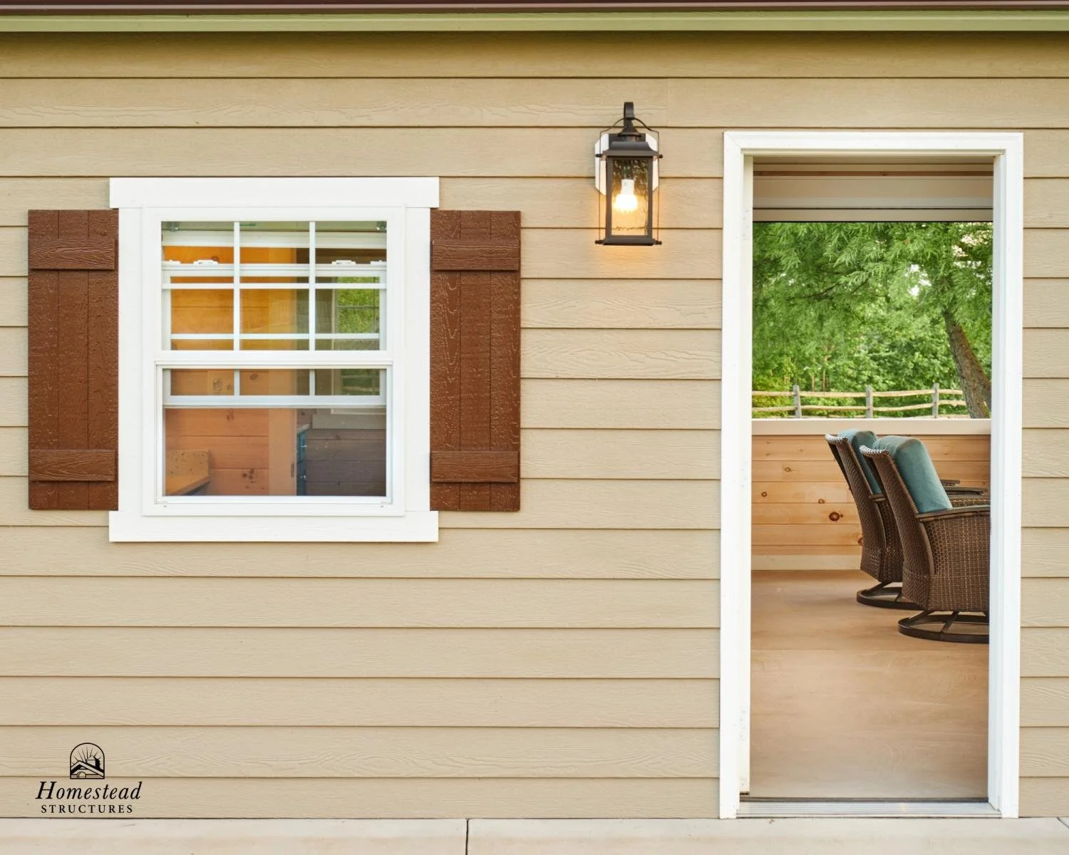 Exterior view of a house showing a beige horizontal siding wall, a white-framed window with brown shutters, a black outdoor lantern, and an open door leading to a porch with seating and green trees outside.