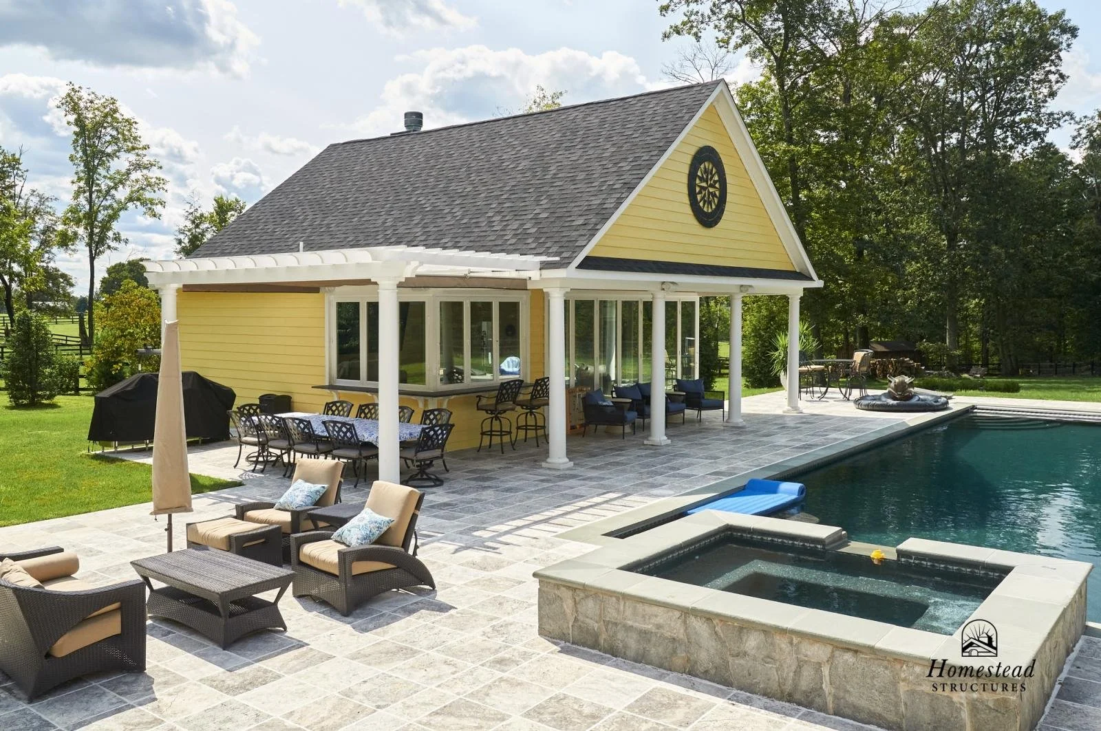 A backyard with a swimming pool and hot tub adjacent to a yellow house with white columns and a shaded patio area, surrounded by greenery and trees.