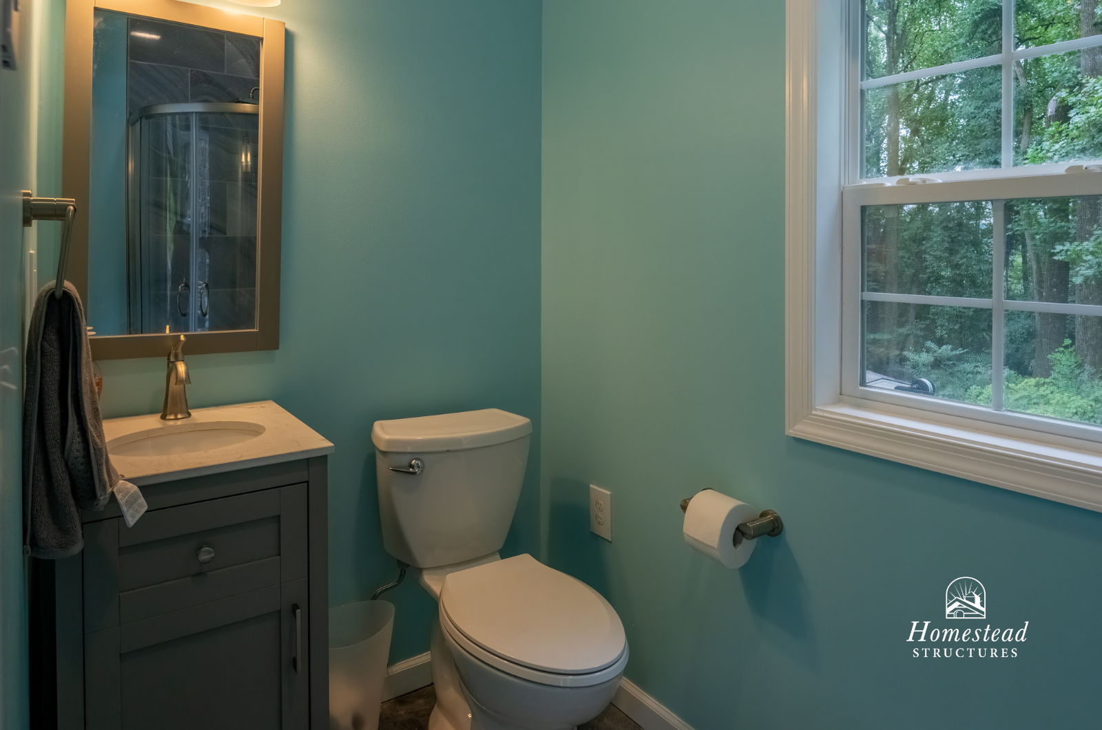 Small bathroom with a toilet, a vanity with a sink and mirror, and a window showing green trees outside.
