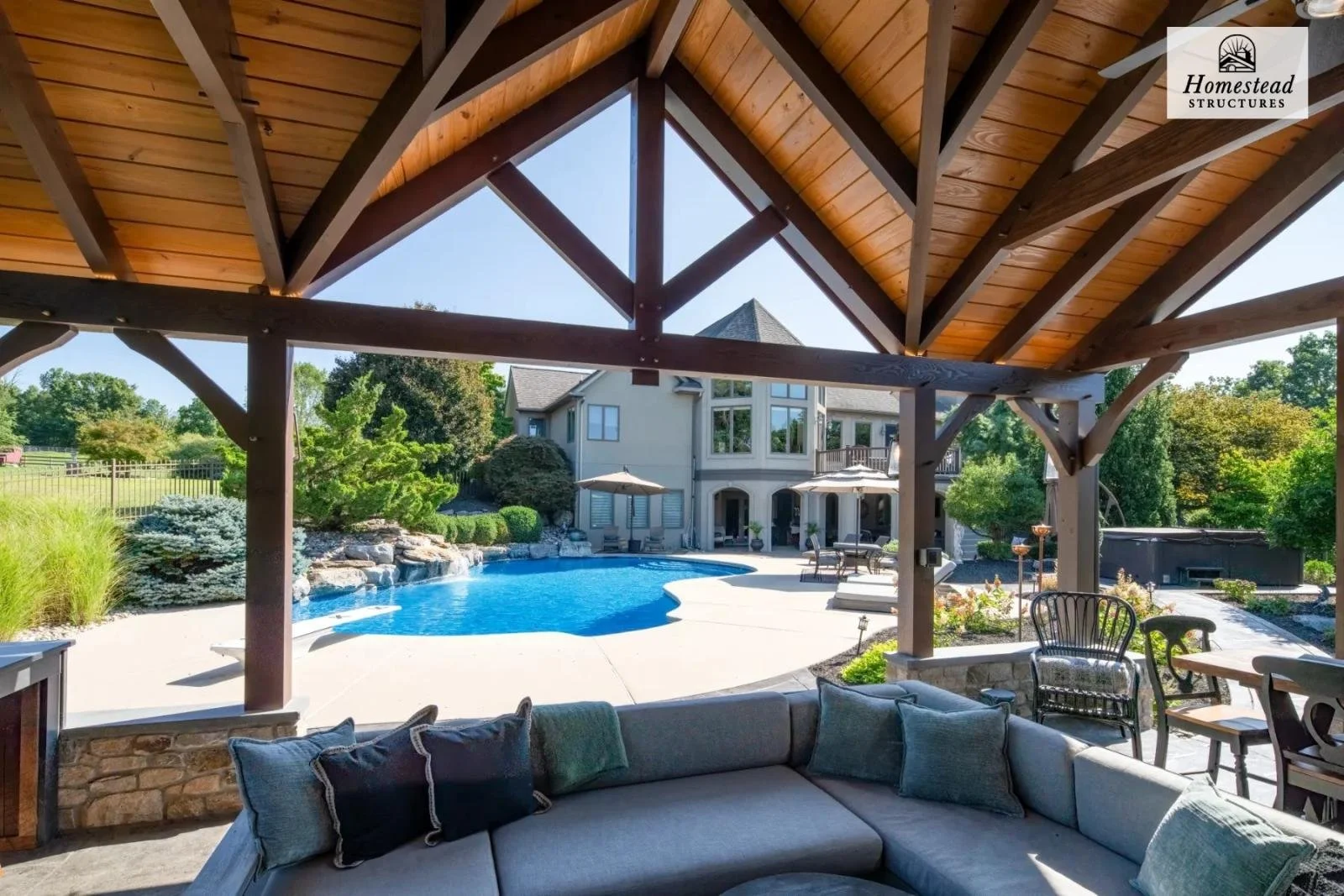 View from covered patio with wooden ceiling and outdoor seating overlooking a backyard swimming pool, house, and lush greenery.