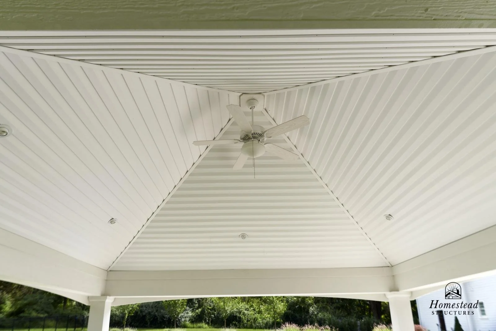 Ceiling fan on a covered porch with white beadboard ceiling and support columns, overlooking a green yard.
