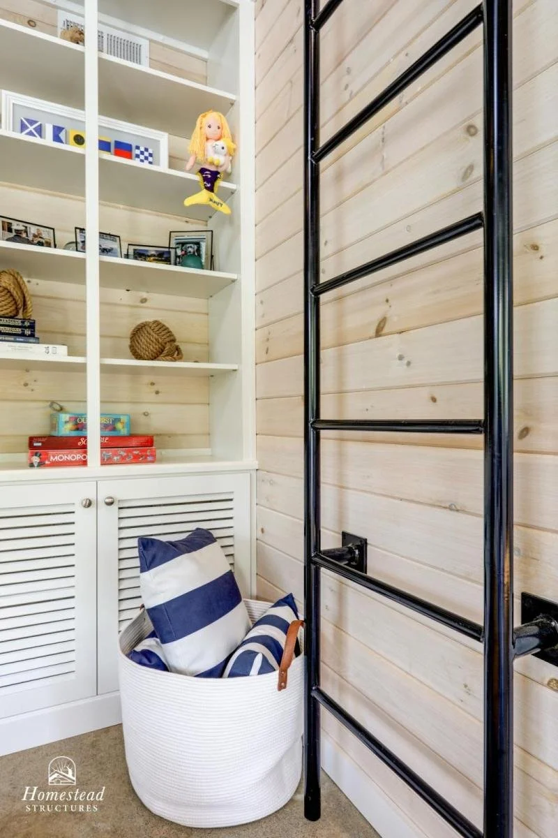 A corner of a room with a white bookshelf holding various books, toys, and photos. There is a plush doll hanging on the shelf. Next to it, a black metal ladder is mounted on a wooden wall. In front of the wall, a white basket contains striped cushion