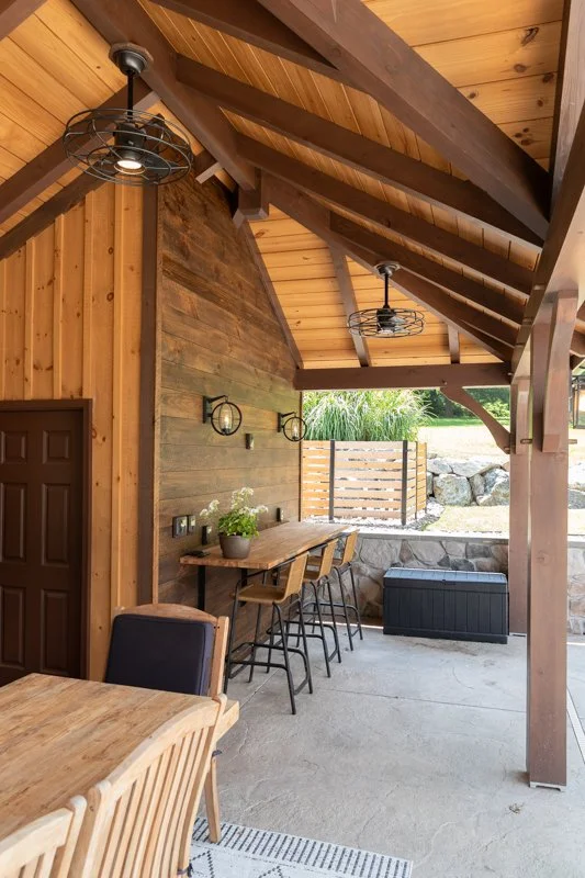 Covered patio area with wooden ceiling and walls, featuring two black industrial-style ceiling lights, a wooden bar table with four chairs, a potted plant, and a view of a yard with rocks and greenery.